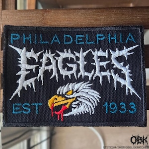 Può includere: Una toppa rettangolare nera con le parole "PHILADELPHIA EAGLES" in lettere bianche e appuntite e testo blu acqua. Sotto, una testa d'aquila con dettagli gialli, rossi e bianchi. La toppa include anche "EST 1933".