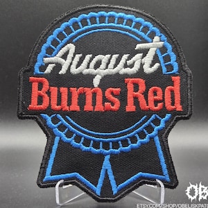 以下が含まれることがあります： 白と赤の「August Burns Red」の文字が刺繍された黒色のパッチ。青と黒のリボンのデザインが施されています。パッチは黒い縁取りがあり、衣類やアクセサリー用である可能性があります。
