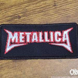 May include: A black rectangular patch featuring the word "METALLICA" in white embroidery with red detailing. The patch is bordered in black and is displayed on a wooden surface. This is a music-themed item.