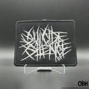 Könnte beinhalten: Schwarzer rechteckiger Aufnäher mit weißem, abstraktem, stacheligem Design. Der Aufnäher ist mit einem komplexen, ineinander verwobenen Muster bestickt. Der Aufnäher wird auf einem transparenten Ständer präsentiert. Die Etsy-Shop-URL ist unten rechts sichtbar.