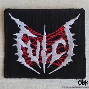 Puede incluir: Parche cuadrado negro con un diseño bordado en blanco y rojo. El diseño presenta texto estilizado con elementos angulares y afilados. El parche tiene un borde y es adecuado para coser o planchar sobre tela. El texto está parcialmente oscurecido por el diseño.