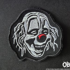 May include: An embroidered patch featuring a black background with a white-stitched clown face design. The clown has messy hair, dark eye makeup, and a prominent red nose. The patch has a black stitched border and a jagged, irregular shape.
