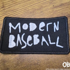 Könnte beinhalten: Schwarzer rechteckiger Aufnäher mit den weißen Worten "MODERN BASEBALL". Der Aufnäher hat einen schwarzen Rand und liegt auf einer Holzoberfläche. Es ist ein Musik-Themenartikel.