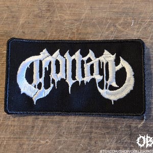 May include: A black rectangular patch with white embroidered text. The text is stylized and appears to be a band logo. The patch has a black border. The bottom right corner has the text "OBK" and a website address.