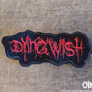 May include: A black embroidered patch with a jagged, irregular border. The text DYING WISH is stitched in a bright red, stylized, distressed font that resembles dripping paint or ink, featuring sharp, elongated lines and a textured appearance.