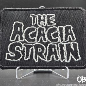 May include: A black rectangular patch with white embroidered text reading "THE ACACIA STRAIN." The patch has a black border and is displayed on a clear stand. A white logo and Etsy shop URL are in the bottom right corner.