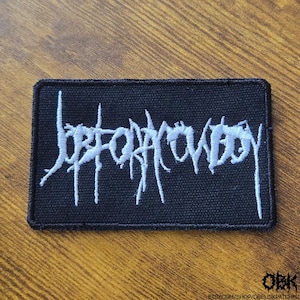 Può includere: Una toppa rettangolare nera con la scritta bianca ricamata "Jefecowboy" in un carattere gotico. La toppa è progettata per essere applicata su abbigliamento o accessori.