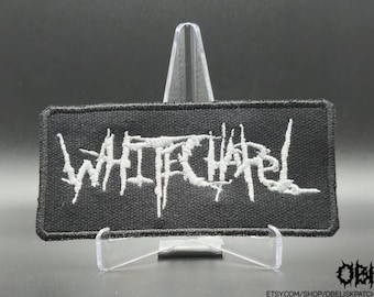 May include: A rectangular black embroidered patch featuring the word Whitechapel in a stylised, jagged white font. The patch has a thick black border and is displayed on a clear plastic stand against a dark background, with small text in the corner.