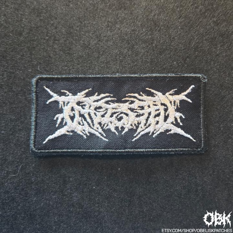 May include: A rectangular black embroidered patch featuring white thread in a stylised, jagged, and intricate lettering design. The embroidery has a dense texture with sharp, pointed edges throughout the central text graphic on a black fabric base.