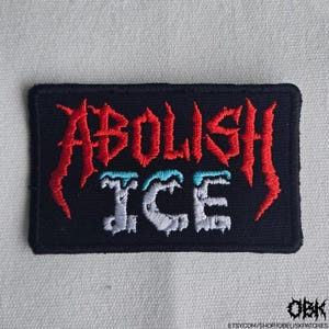 May include: Black rectangular embroidered patch with the words "ABOLISH ICE" in red, light blue, and white thread. The patch has a stitched border and the text is in a stylized font.