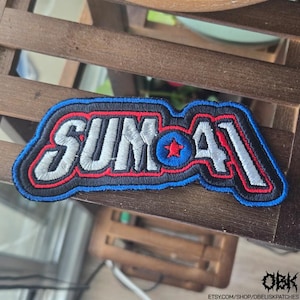 Puede incluir: Parche bordado con el texto "SUM 41" en blanco, contorneado en rojo y negro, con un borde azul. Una estrella roja está centrada en la "O". El parche rectangular es para ropa o accesorios.