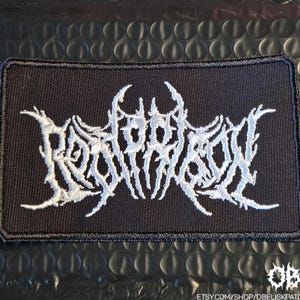 May include: A rectangular black embroidered patch featuring white text in a stylized, jagged, and intricate death metal font. The embroidery has a dense, textured appearance with a thick black border surrounding the central white lettering.
