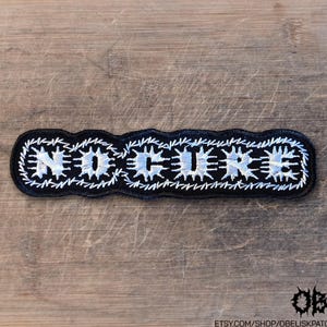 May include: A rectangular embroidered patch with a black background and white stitched border. The text NO CURE is centered in white, stylized lettering with a jagged, spiked appearance. The patch features a thick, textured white thread outline.