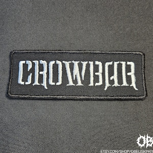 May include: Black rectangular patch with the word "CROWBAR" embroidered in white gothic lettering. The patch is on a dark gray textured background. The bottom right corner has the text "OBK" and a website address.