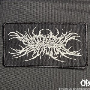 Könnte beinhalten: Ein rechteckiger, schwarz bestickter Aufnäher mit einem weißen, komplexen und gezackten symmetrischen Design, das an stilisierte, dornige oder wurzelartige Schriftzüge erinnert. Der Aufnäher hat einen gestickten Rand auf dunklem Stoff.