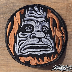 May include: A circular embroidered patch featuring a stylized silver stone-like face with a downturned mouth centered on a black background, surrounded by orange flame-like shapes, with a black stitched border and a metallic-style logo in the corner.