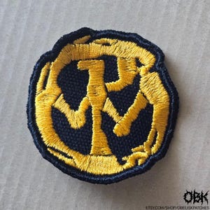 May include: Embroidered patch with a circular design. The patch features a gold-colored symbol on a navy blue background. The symbol appears to be a stylized letter or emblem. The patch has a black border and is likely for clothing or accessories.