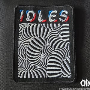 May include: Embroidered patch with the word "IDLES" in red, white, and blue lettering. The patch features a black background with a swirling, black and white striped design. The patch is rectangular.