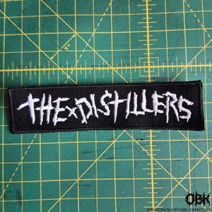 May include: Black rectangular patch with the white text "THE DISTILLERS". The patch is embroidered with a distressed font. The patch is on a green cutting mat.