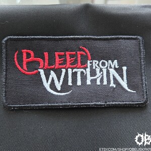 May include: A rectangular black embroidered patch featuring the text BLEED in red thread and FROM WITHIN in white thread. The patch has a black stitched border and is attached to a black fabric surface with a visible weave texture.