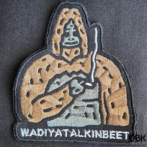 Op de afbeelding: Geborduurde patch met een figuur met een capuchon die een staf vasthoudt. De patch heeft een zwarte rand, met details van beige, grijs en wit garen. De tekst "WADIYATALKINBEET" is onderaan geborduurd. Uniek ontwerp.