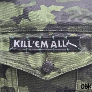 May include: Close-up of a green and black camouflage jacket with a black patch that reads "KILL'EM ALL" in white lettering. The patch is stitched with green thread. A silver button is visible.