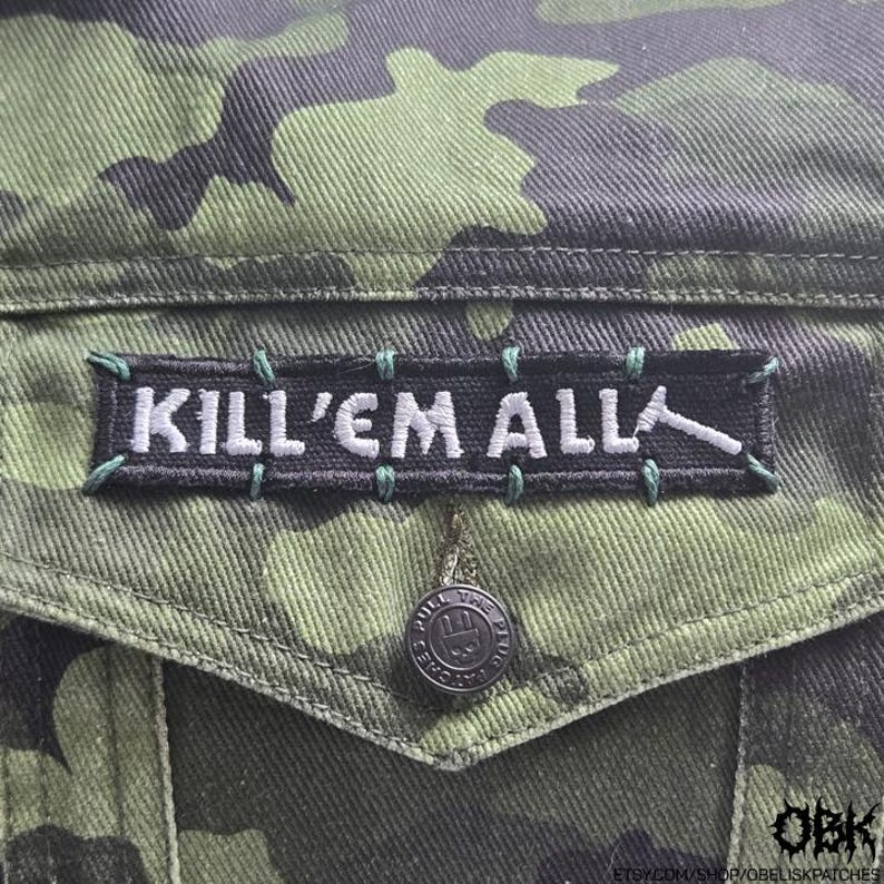 Puede incluir: Primer plano de una chaqueta de camuflaje verde y negra con un parche negro que dice "KILL'EM ALL" en letras blancas. El parche est&aacute; cosido con hilo verde. Se ve un bot&oacute;n plateado.