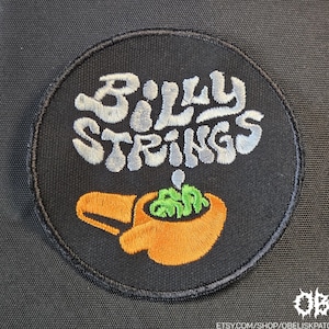 Può includere: Toppa ricamata circolare nera con la scritta "Billy Strings" in bianco e azzurro, sopra un disegno arancione e verde. L'URL del negozio Etsy è visibile in basso.