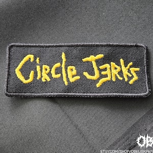 May include: Black rectangular patch with yellow embroidered text that reads "Circle Jerks". The patch is on a black fabric background. The patch is a rectangle with rounded corners.