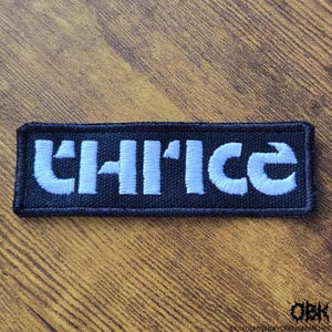 May include: A rectangular black patch with the word "THRICE" embroidered in white, block letters. The patch has a black border and is likely designed to be sewn or ironed onto clothing or accessories. The patch is on a wooden surface.