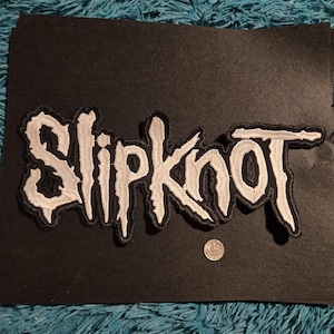 May include: Embroidered patch featuring the band name "Slipknot" in a white, stylized font with a black outline. The patch is on a black background, with a quarter coin for size reference. The patch is likely for a jacket or bag.