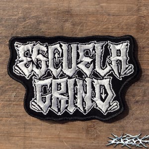 May include: A black embroidered patch featuring the white text ESCUELA GRIND in a stylised, jagged font. The patch has a thick black border and a textured fabric base, with the letters arranged in two lines against the dark background.