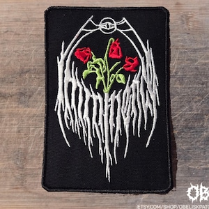 May include: A rectangular black embroidered patch featuring a white stylized graphic of a central eye within an arch, surrounded by hanging white tendrils and three red roses with green stems and leaves. The design is centered on the black fabric.
