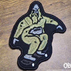 Könnte beinhalten: Ein gestickter Aufnäher, der eine hellgrüne Kreatur mit Bart und Hut auf einem Skateboard zeigt. Das Motiv ist mit schwarzem Garn umrandet und hat hellblaue Akzente an Händen, Füßen und Skateboardrollen. Der Text OBK ist zu sehen.
