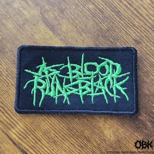 May include: Black rectangular patch with green embroidered text that reads "BLOOD IN BLACK". The patch has a black border and is likely for clothing or accessories.