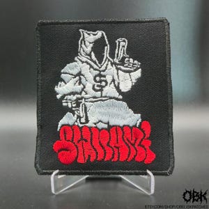 May include: Square black patch with a white hooded figure holding a gun, with the word "SHARK" in red. The patch is embroidered with detailed stitching and is displayed on a clear stand.