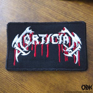 May include: Black rectangular patch with the word "Mortician" in white, stylized lettering. Red drips extend from the letters. White, claw-like designs frame the text. The patch is on a wooden surface.