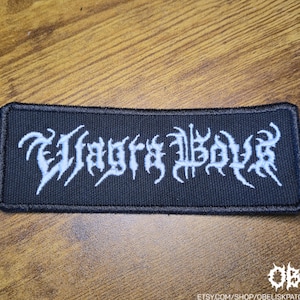 Peut inclure: Un écusson rectangulaire noir avec des lettres gothiques blanches qui se lisent "Wagrs Boys". L'écusson est probablement destiné aux vêtements ou aux accessoires. L'écusson est sur une surface en bois.