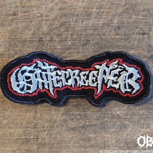 May include: A black embroidered patch featuring the word GATECREEPER in stylized, jagged silver lettering outlined in red. The patch has a distinct, irregular shape that follows the contours of the text, with a black border surrounding the design.