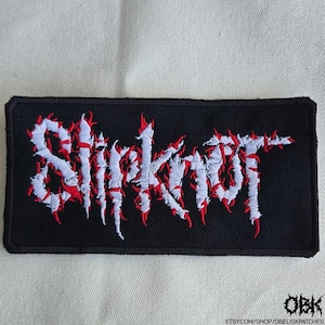 May include: Black rectangular patch with the word "Slipknot" embroidered in white and red. The text has a jagged, flame-like design. The patch is likely for clothing or accessories.