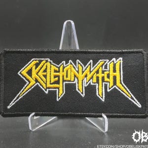 Puede incluir: Parche rectangular negro con el texto amarillo "SKELETONWITCH" contorneado en blanco. El parche se muestra sobre un soporte de plástico transparente sobre un fondo oscuro. El parche es probablemente para una chaqueta o bolso.