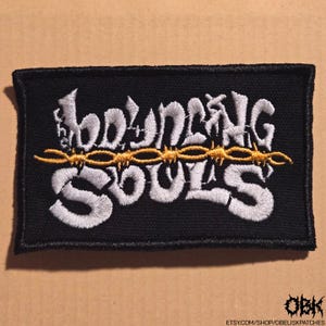 May include: Black rectangular patch with the words "BOUNCING SOULS" embroidered in white, with a gold barbed wire design. The patch has a black border and the text "OBK" and a website address at the bottom.