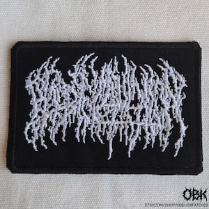 Può includere: Toppa rettangolare nera con testo ricamato bianco. Il testo sembra essere un logo o un design di una band, con un carattere stilizzato e astratto. La toppa ha un bordo nero ed è probabilmente in tessuto.