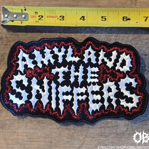 May include: A black embroidered patch with white text reading AMYL AND THE SNIFFERS, featuring red stitching accents around the letters. A yellow measuring tape is positioned above the patch, showing a width of approximately 7.5 inches.