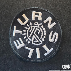 Könnte beinhalten: Ein runder, schwarzer Aufnäher mit weiß gesticktem Text und einem zentralen Design. Der Text "URNS TILE" ist am Rand angeordnet. Das zentrale Design zeigt stilisierte Formen und Linien. Der Aufnäher ist für Kleidung oder Accessoires.
