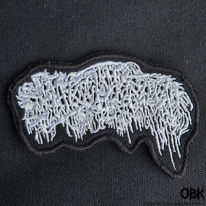 Może przedstawiać: Haftowana naszywka z czarną obwódką i białym, stylizowanym wzorem. Wzór zawiera nieczytelny tekst w kształcie kropli, tworząc gotycką lub metalową estetykę. Naszywka jest prawdopodobnie przeznaczona na odzież lub akcesoria.