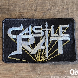 May include: Black rectangular patch with the text "CASTLE RAT" in white, stylized lettering. A sword design is centered within the text. Yellow rays emanate from behind the text. The patch is embroidered.