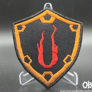 May include: Embroidered shield patch with a black background, orange and gold border, and a red flame design. The patch is displayed on a clear stand. The Etsy shop name is visible in the bottom right corner.