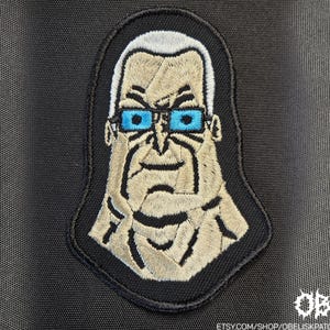 Op de afbeelding: Een geborduurde patch met een gestileerd personage met wit haar, felblauwe rechthoekige bril en een gebruinde teint. Het ontwerp is omlijnd met zwart garen op een zwarte stoffen achtergrond, met de tekst OBK zichtbaar in de hoek.