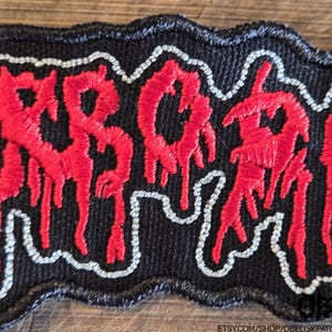 Puede incluir: Un parche bordado rectangular con fondo negro y letras rojas estilizadas que parecen gotear. El texto est&aacute; delineado con un hilo blanco fino, y todo el parche tiene un borde grueso cosido en negro con un contorno ondulado.